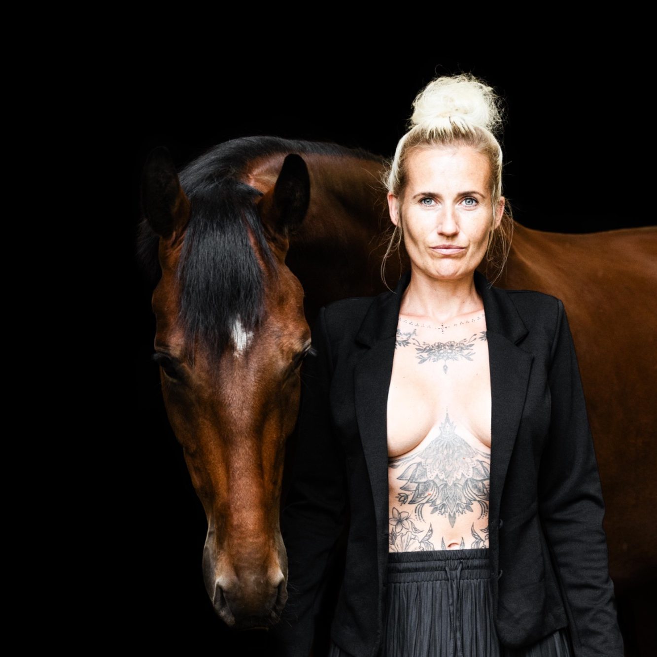 Frau mit tätowiertem Oberkörper steht neben einem Pferd vor schwarzem Hintergrund.